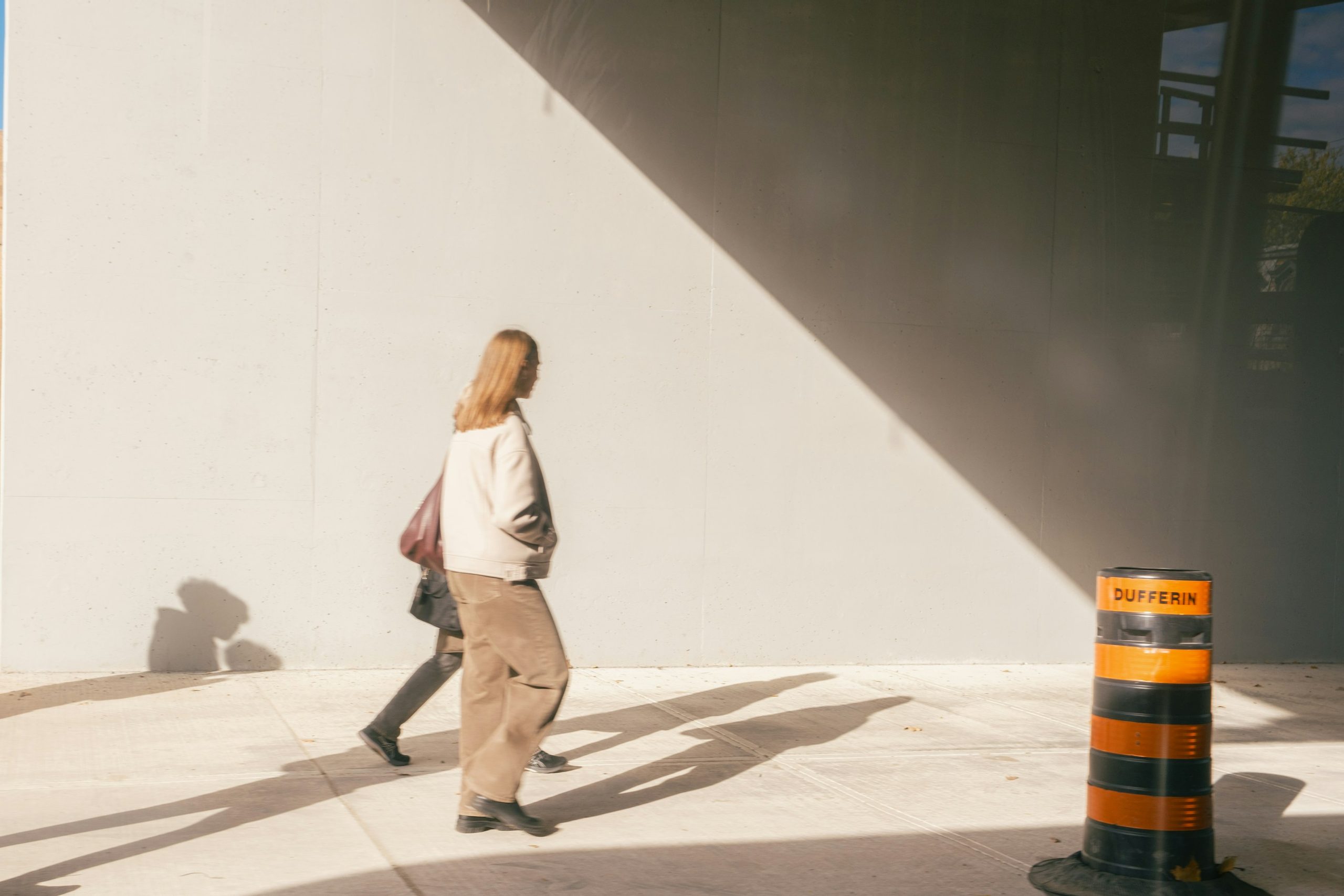 Two pedestrians cast long shadows beside a dark tiled wall.