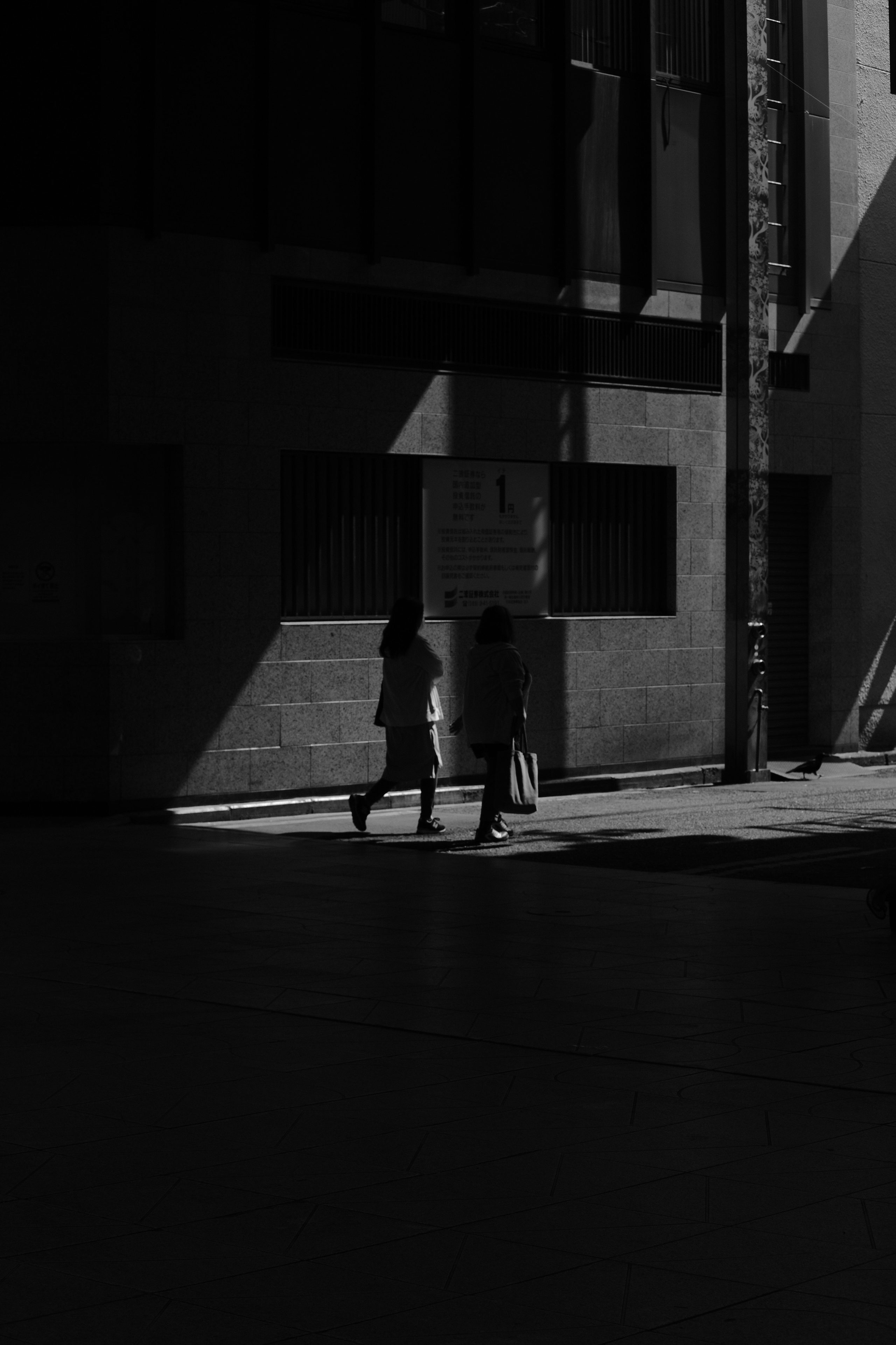 Two people walk beside a tall stone building through angled sunlight.
